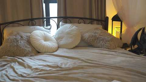 Media: Video of a cozy bedroom with a wrought-iron bed, white and cream textured pillows, beige curtains, a glowing lantern, and soft, warm lighting.