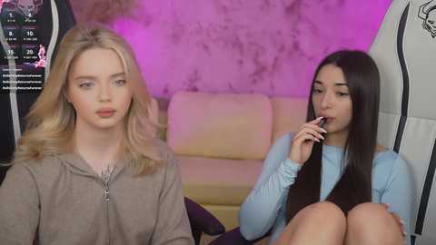 Media: Video of two young women in a modern, dimly lit room. Blonde woman with wavy hair in brown hoodie sits on left, dark-haired woman in blue top and black skirt holds lip gloss, sitting on right.