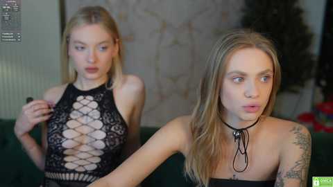 Media: Video of two young women, one with blonde hair, wearing a black lace dress, the other with a tattooed arm, sitting on a green couch in a dimly lit room.