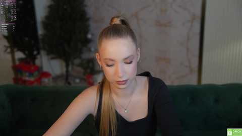 Media: Video of a fair-skinned, slim young woman with blonde hair in a high ponytail, wearing a black long-sleeve top, seated on a green velvet sofa. Background includes a decorative screen and a Christmas tree.