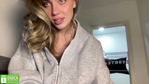 Media: A video of a young Caucasian woman with wavy, blonde hair, light skin, and light makeup, wearing a grey hoodie, standing indoors with a white door and clothes hanging in the background.