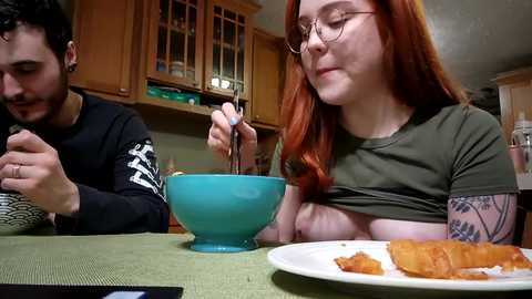 Media: Video of a young couple eating in a cozy kitchen; man with short dark hair, glasses, black shirt, fork in hand; woman with long red hair, olive-green shirt, tattoos, eating pizza.