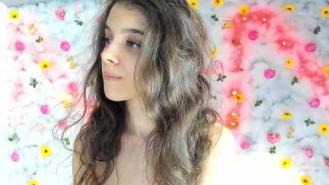 Media: Video of a young woman with long, wavy brown hair, fair skin, and a contemplative expression. She stands against a backdrop of pink and yellow flowers, creating a whimsical, light-hearted atmosphere.