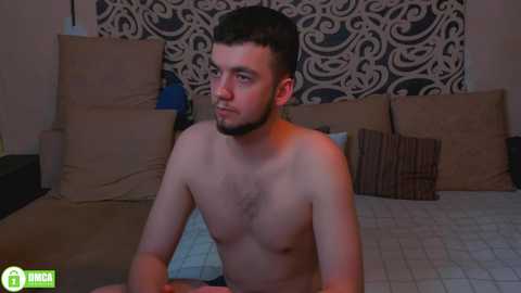 Media: Video of a shirtless, muscular young man with a beard, sitting on a bed with beige pillows and a patterned wall in the background.