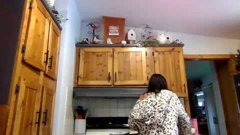 Media: Video of a woman with shoulder-length brown hair in leopard-print pajamas standing in a cozy wooden kitchen with open cabinets, white tiles, and bird-themed decor.