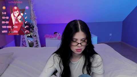 Media: Video of a young woman with long black hair, glasses, and a white sweater, sitting on a bed in a dimly lit room with blue lighting and a TV displaying a game character.