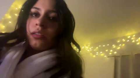 Video of a young woman with long black hair, wearing a white robe, in a dimly lit room with yellow fairy lights and a cream-colored curtain.