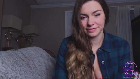 Media: Video of a smiling, fair-skinned woman with long, wavy brown hair, wearing a blue plaid shirt, sitting on a gray bed with a lamp and a plant in the background.