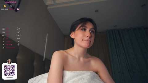 Media: Video of a young woman with fair skin and dark hair, wrapped in a white towel, in a dimly lit bedroom.