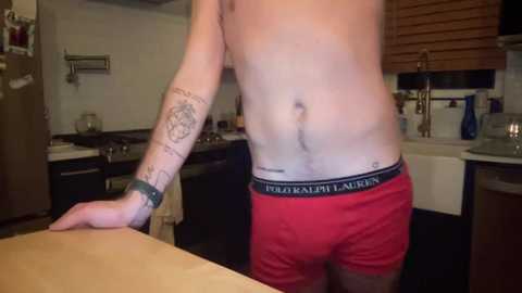 Media: Video of a shirtless man in red Calvin Klein underwear, with tattoos on his arm and back, standing in a dimly lit kitchen.