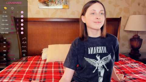 Media: Video of a young Caucasian woman with light skin and straight brown hair, wearing a black Nirvana t-shirt, sitting on a bed with a red plaid blanket. Background shows a wooden headboard, a nightstand with a lamp, and a patterned wallpaper.