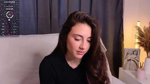 Media: Video of a young Caucasian woman with long brown hair, wearing a black top, sitting on a beige couch in a dimly lit room with a star chart poster, a candle, and a decorative plant in the background.