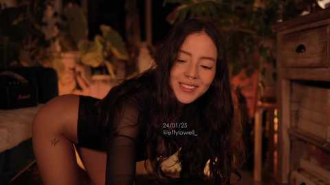 Media: Video of a smiling, dark-skinned woman with long, dark hair, wearing sheer black lingerie, in a dimly lit, tropical-themed room with plants and wooden furniture.