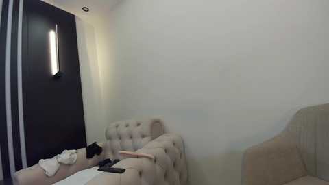 Media: A video of a minimalist, modern bedroom with beige walls and a tufted beige headboard. A white shirt and black pants are folded on a chair. A large dark wooden door with vertical slats and a bright lamp are visible.