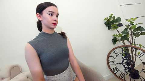 Media: Video of a young woman with light skin and dark hair in a braided ponytail, wearing a grey sleeveless top and white shorts, standing indoors next to a large plant in a white room.