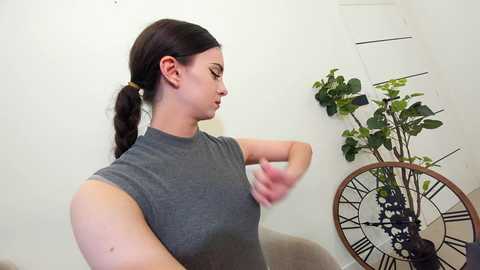 Media: A video of a young woman with a ponytail, wearing a grey sleeveless shirt, adjusting her bra. She stands next to a clock with greenery in a white room.