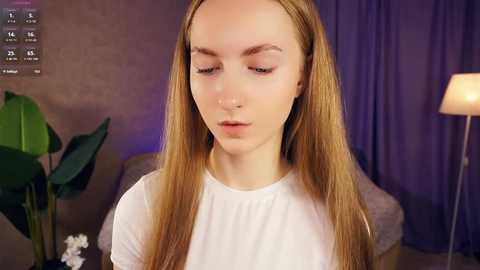 Video of a young Caucasian woman with fair skin and long, straight, light brown hair, wearing a white blouse. She has a contemplative expression. Background features a green plant, purple curtains, and a lamp.