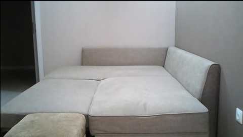 Media: A video of a minimalist, modern living room corner with a beige sectional sofa against a plain, light gray wall, featuring a cream-colored cushioned backrest and armrests. The background includes a dark wooden door frame and a beige carpeted floor.