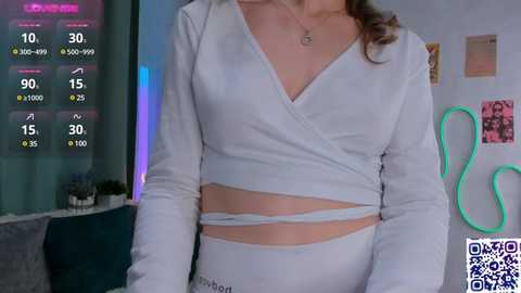 Media: Video of a woman with light skin and medium-length brown hair, wearing a white, long-sleeve crop top and high-waisted white pants, in a room with neon lights and colorful artwork on the wall.