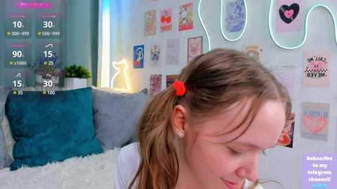 Media: A video of a young, fair-skinned girl with light brown hair in a ponytail, wearing a white top and red hair tie, sitting in a cozy, brightly lit room with blue and white decor, various posters, and a neon sign.