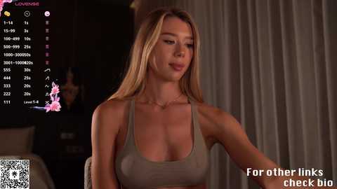 Media: Video of a young, blonde woman with long hair, wearing a revealing, olive-green tank top that highlights her medium-sized breasts, sitting indoors. The background includes a dimly lit room with a window, a black screen displaying stats, and a QR code.