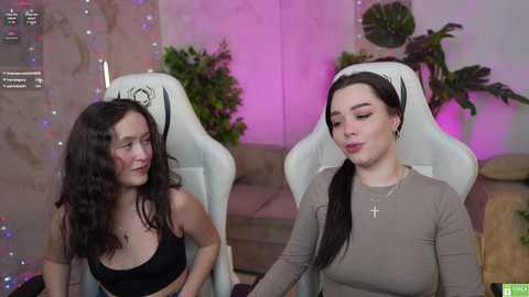 Media: Video of two women in a modern, pink-lit room. One, with long dark hair, wears a black top; the other, with long brown hair, wears a beige top. Both seated in white gaming chairs.