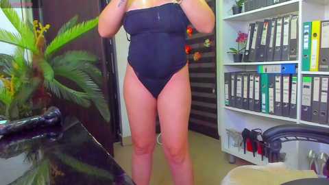 Media: Video of a woman with light skin and a full figure wearing a black one-piece swimsuit, standing in a modern office with a potted palm, bookshelves, and a black desk.