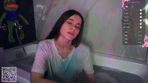 Media: Video of a young woman with long brown hair, wearing a white t-shirt, in a bathtub, surrounded by colorful, multicolored lights.