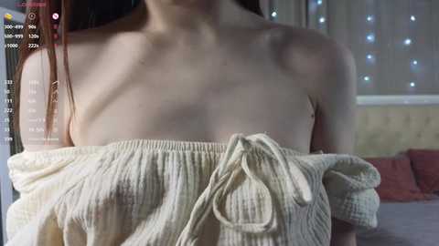 Media: A video of a fair-skinned woman with long red hair, wearing a white off-shoulder top, partially revealing her small breasts, in a dimly lit room with a bed and string lights in the background.