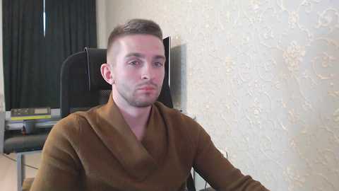 Media: Video of a young man with short brown hair, wearing a brown sweater, sitting in a black office chair against a textured cream wall with a TV on the left.