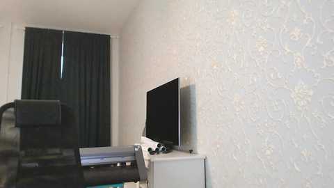 Media: Video of a minimalist room with light beige floral wallpaper, black curtains, a black office chair, a white desk, and a black flat-screen TV mounted on the wall.