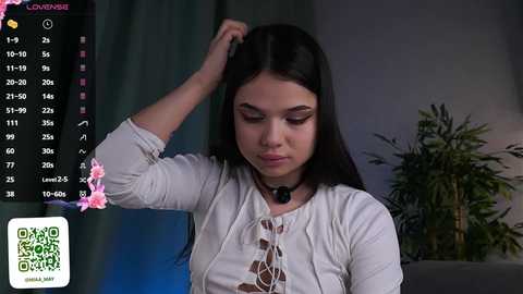 Media: Video of a young woman with long black hair, wearing a white top, seated indoors with a green plant in the background.