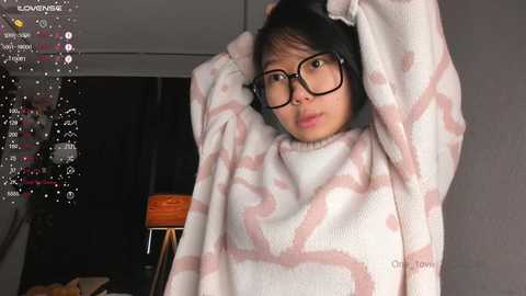 Media: Video of an Asian woman with medium skin tone, wearing black-rimmed glasses and a white, pink-patterned sweater, adjusting her headphones. Background includes a dimly lit room with a wooden stool and a partially visible person.