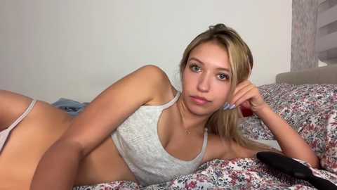 Media: Video of a blonde woman with fair skin, medium build, lying on a floral-patterned bed in a casual grey sports bra and panties, looking relaxed.