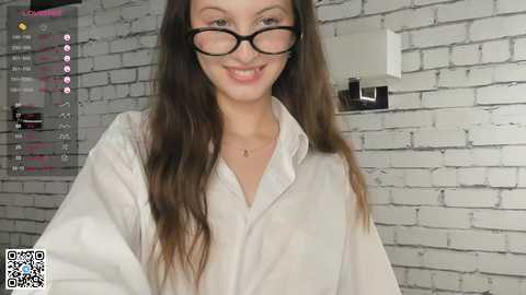 A video of a smiling young woman with long brown hair, wearing black-rimmed glasses, a white lab coat, and standing in front of a white brick wall.