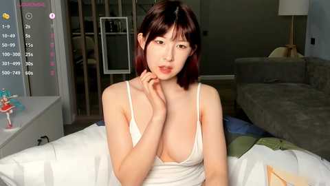 A video of a young Asian woman with straight, shoulder-length brown hair, wearing a white camisole, sitting on a couch in a modern living room.