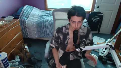 Media: A video of a young woman with short dark hair, wearing a partially unbuttoned black and white shirt, sitting on a blue carpet in a messy bedroom, recording a video with a white microphone.