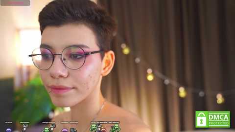 Video of a young Asian woman with short, dark hair, wearing glasses and a pink top, smiling. Background features soft-focus curtains and string lights. \"DMCA Protected\" watermark in the corner.