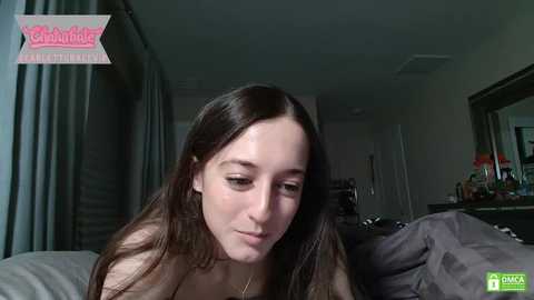 Media: A candid video of a young woman with long brown hair, wearing a black top, sitting on a bed in a dimly lit bedroom.