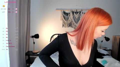 Media: Video of a woman with shoulder-length, straight red hair, wearing a black, low-cut top, and a tattoo on her right shoulder, in a modern bedroom with a black and white macram\u00e9 wall hanging.