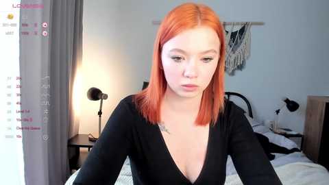 Media: Video of a pale-skinned woman with shoulder-length red hair, wearing a black V-neck top, sitting on a bed with white sheets in a softly lit bedroom.