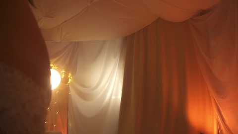 Media: Video of a dimly lit, warm-toned tent with soft, draped fabric. A golden, glowing light source is visible through the fabric, creating a mystical, cozy ambiance.