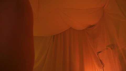 Media: A dimly lit video shows a close-up of a woman's lower abdomen and thighs, partially covered by a sheer, orange fabric. The image has a warm, intimate atmosphere with soft, diffused lighting.