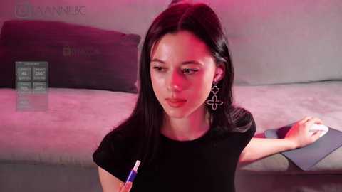 Media: Video of a young woman with long dark hair, wearing a black top, holding a pen, seated on a beige couch in a modern room with red lighting and a digital screen on the left.