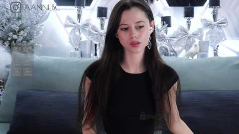 Media: Video of a woman with long, straight black hair, wearing a black dress and silver earrings, sitting on a teal sofa in a modern, chic living room with white drapes and ornate silver chandeliers.