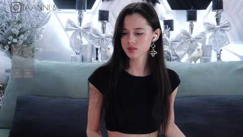 Media: Video of a slender young woman with long black hair, wearing a black crop top and dangling cross earrings, seated on a gray couch in a modern, minimalist room with white decor.