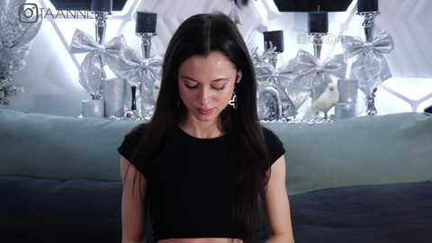 Media: Video of a slender, fair-skinned woman with long, straight black hair, wearing a black crop top, sitting indoors. Background features a festive, metallic, silver and white decor with bows and candles.