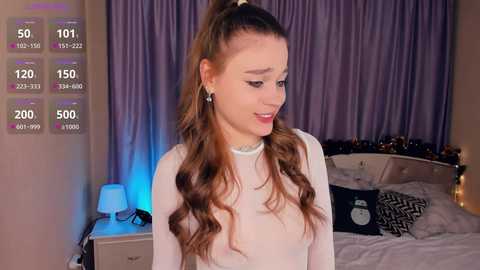 Media: Video of a young woman with long, wavy brown hair in a ponytail, wearing a white long-sleeve top, standing in a bedroom with a purple bedspread, gray curtains, and a nightstand with a lamp.