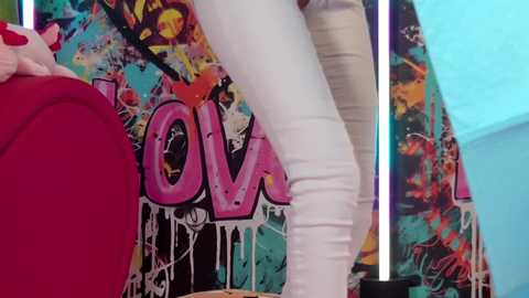 Media: Video of a person in white pants and a red jacket standing near a vibrant, graffiti-covered wall. The background is colorful and textured, suggesting an urban or street art setting.