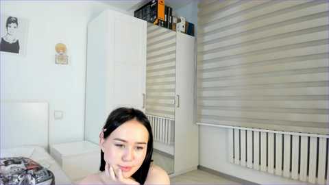 Media: A video of a young Asian woman with long black hair, wearing a white dress, sitting on a bed in a modern, minimalist bedroom with white walls, a radiator, and closed blinds.